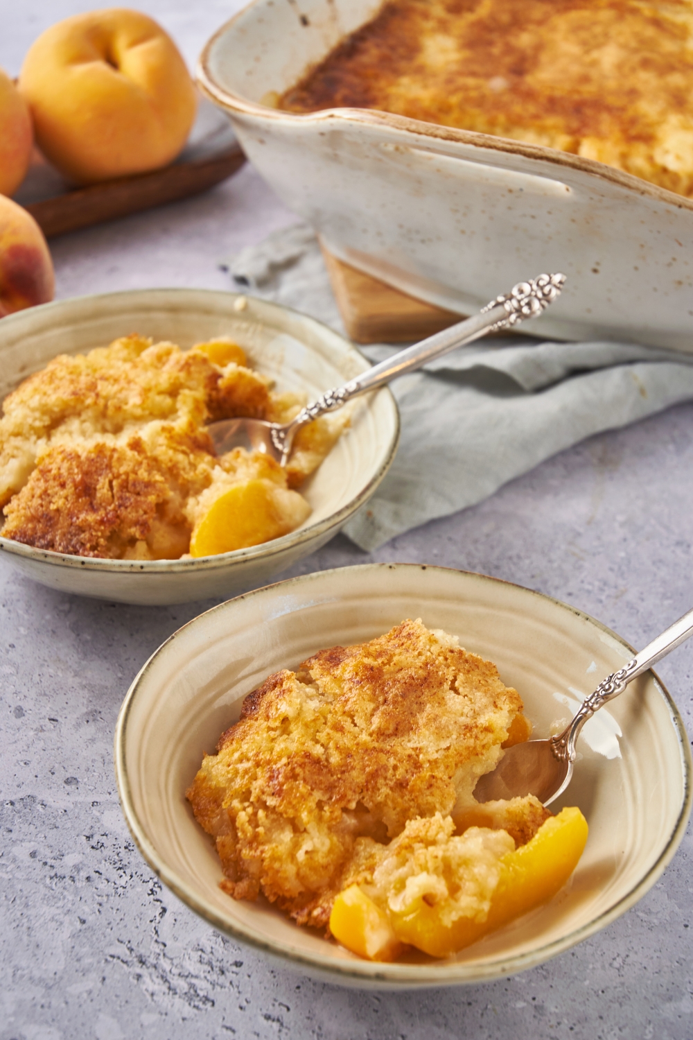 Two servings of peach cobbler with spoons in the bowls and the rest of the cobbler in the background.