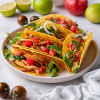 A plate of crispy shredded chicken tacos stacked next to each other with pieces of shredded lettuce, diced tomato, and diced onion in the tacos.