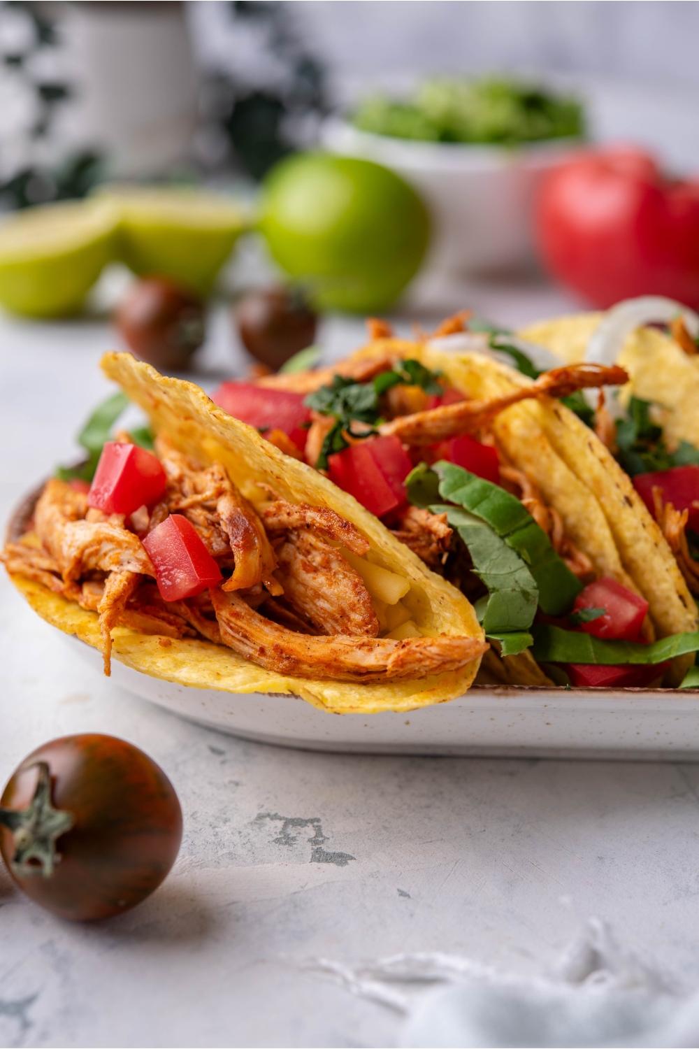 A plate of crispy shredded chicken tacos stacked next to each other with pieces of shredded lettuce, diced tomato, and diced onion in the tacos.