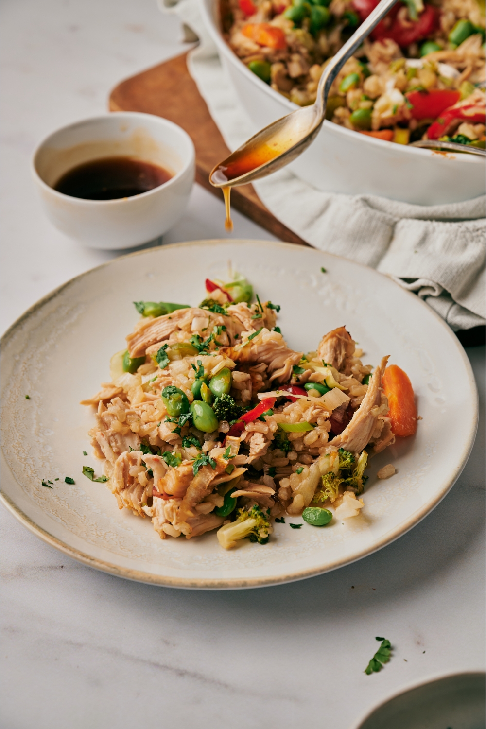 A spoon drizzling sauce over top of a plate of shredded chicken, brown rice, and stir-fried vegetables.