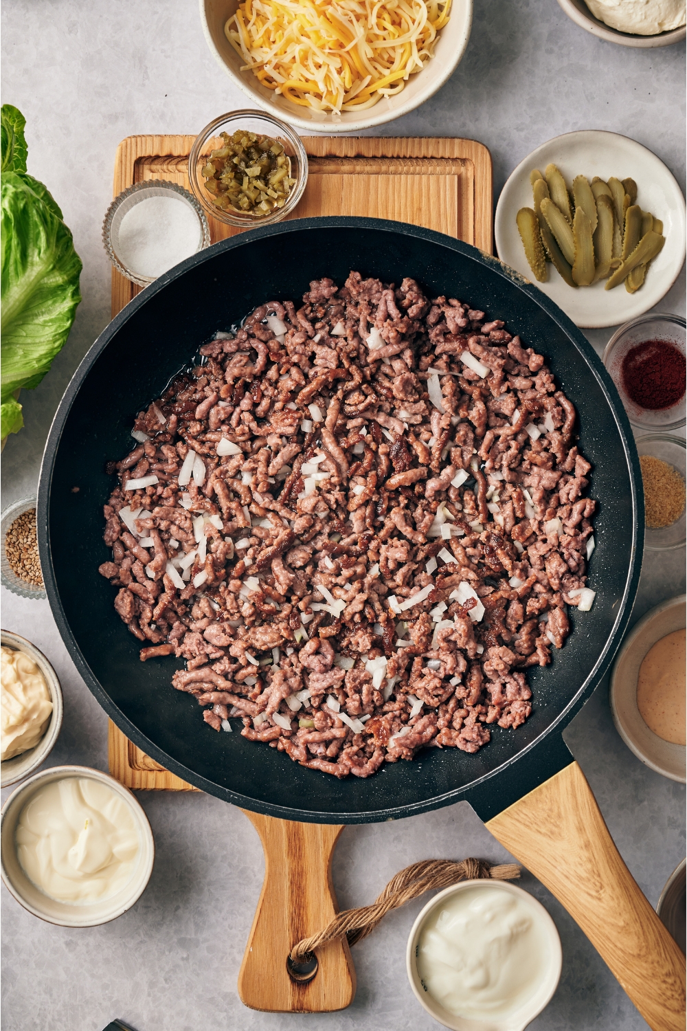 A skillet filled with diced onion and ground beef cooking. The skillet is on a wooden board and surrounded by an assortment of ingredients.