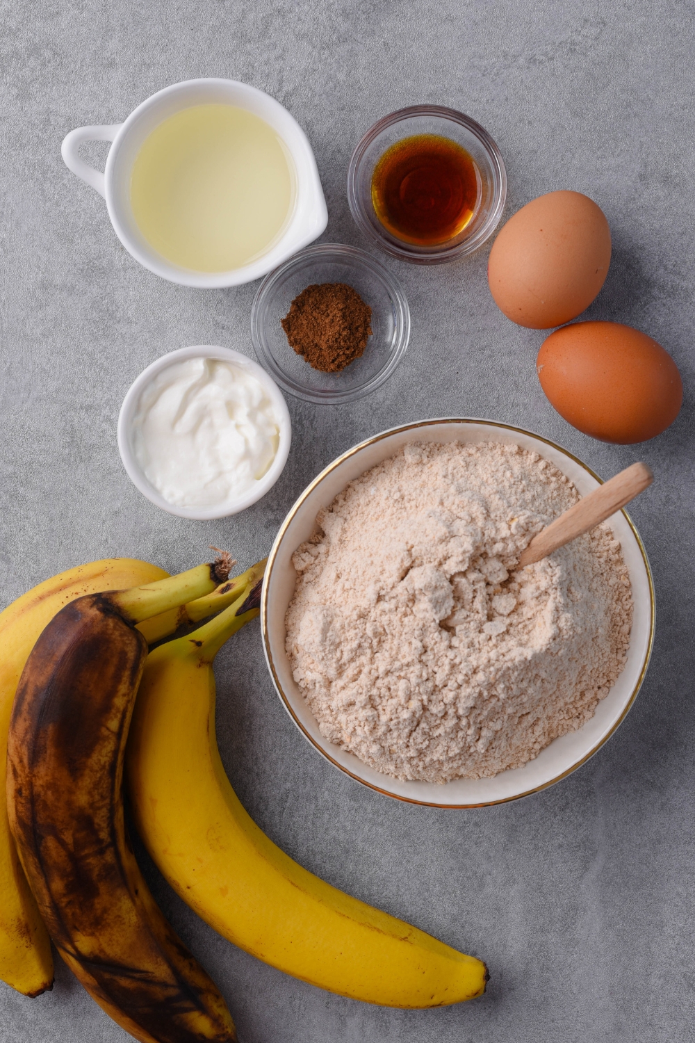 A countertop with bananas, cake mix, sour cream, gingerbread spice, vanilla extract, oil, and eggs laid out in separate bowls and containers.