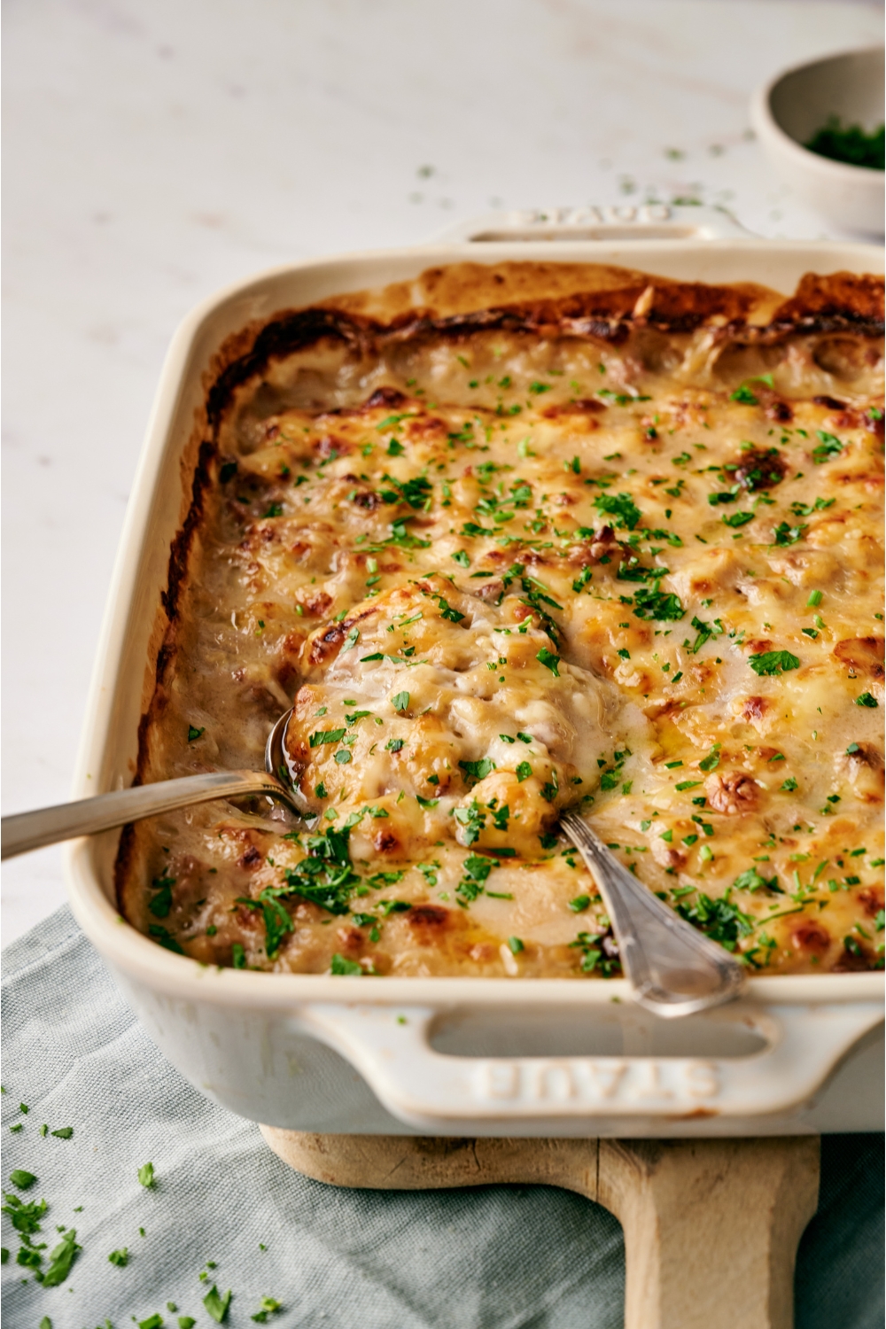 A white baking dish filled with freshly baked casserole covered in a layer of bubbly melted cheese and garnished with fresh herbs. There are two spoons in the casserole.