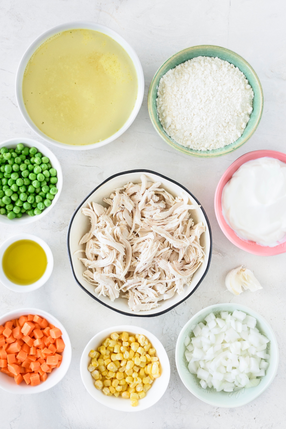 An assortment of ingredients including bowls of cooked and shredded chicken, chicken broth, flour, yogurt, diced onion, diced carrot, corn, peas, and oil.