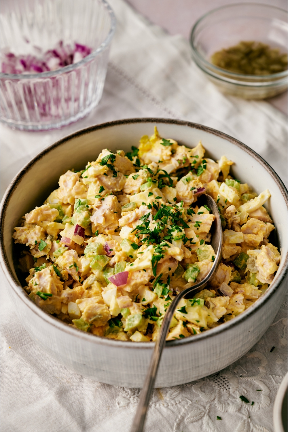 A bowl of chicken and egg salad with pieces of chopped celery, red onion, and fresh green herbs. There is a spoon in the bowl.