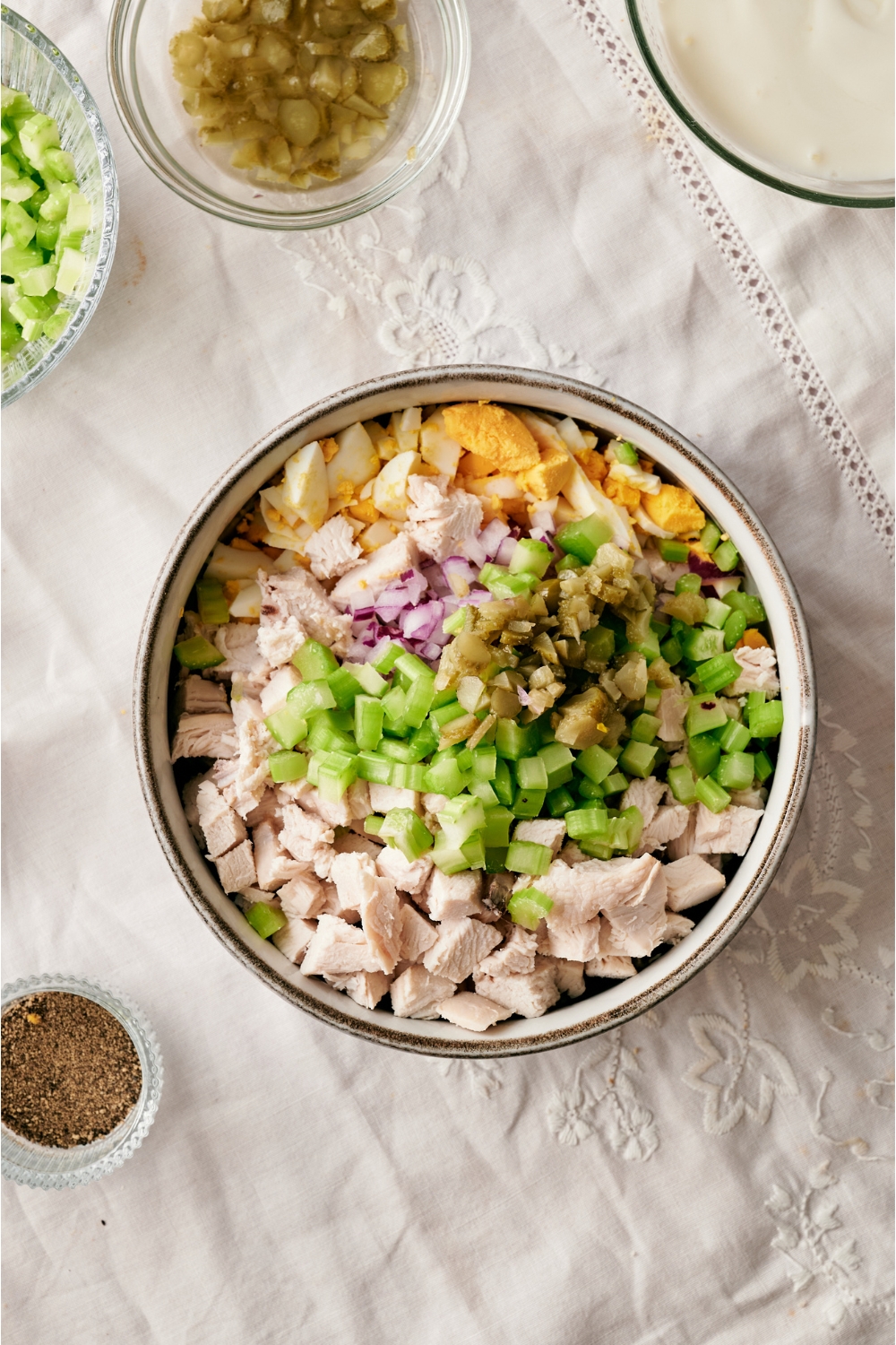 A bowl filled with cubed chicken, diced celery, pickles, red onion, and chopped hard boiled eggs next to smaller bowls of black pepper and chopped pickles.
