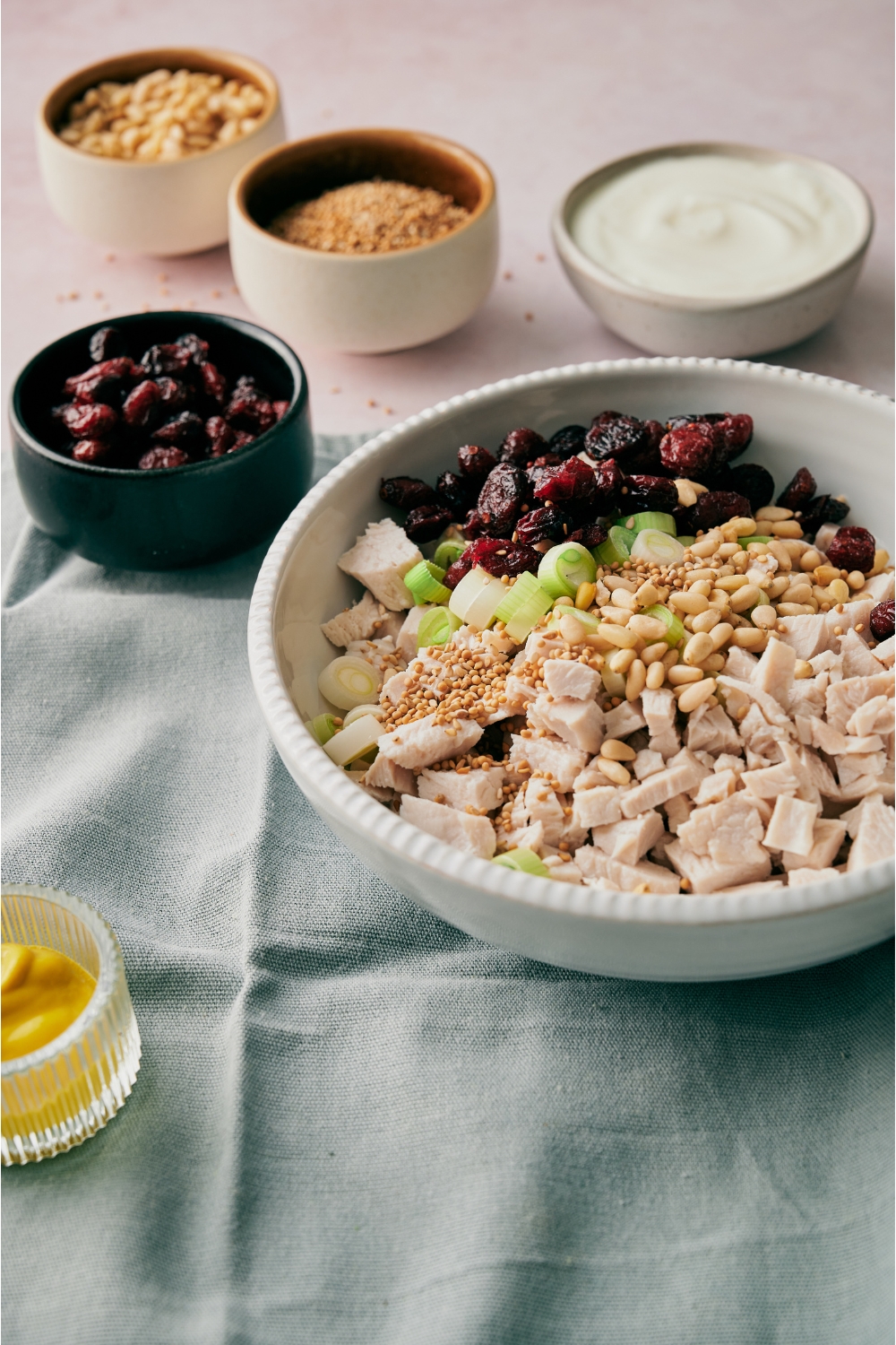A bowl with diced chicken, mustard seeds, pine nuts, sliced green onion, and dried cranberries, next to smaller bowls of mayonnaise, mustard, dried cranberries, mustard seeds, and pine nuts.