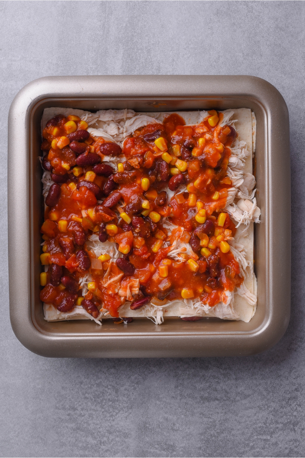 Square baking dish filled with layers of halved flour tortillas, shredded chicken, and a bean, corn, and tomato sauce mixture.