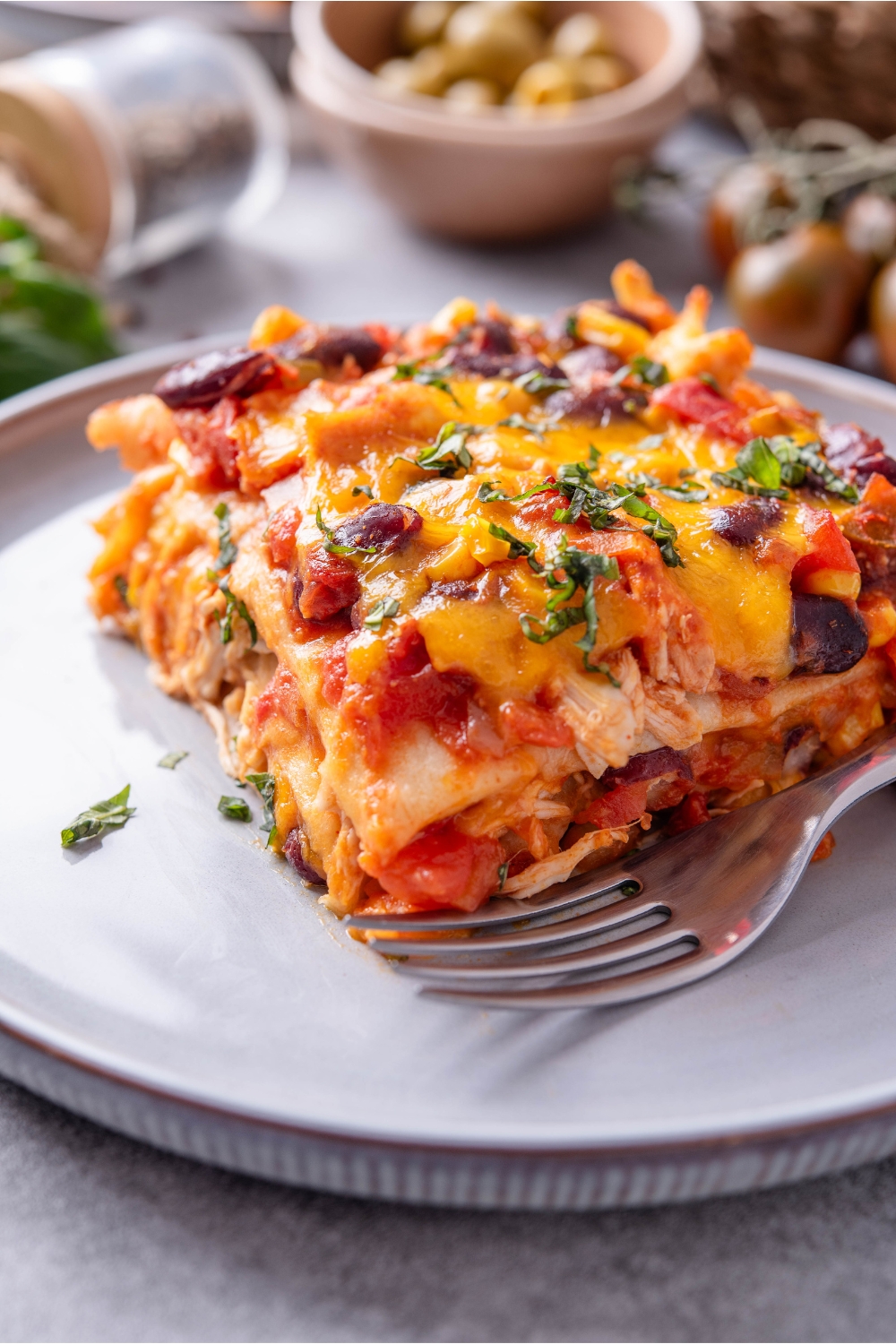 A square serving of chicken tortilla casserole layered with tomato sauce, shredded chicken, black beans, and topped with melted cheese, and fresh green herbs.