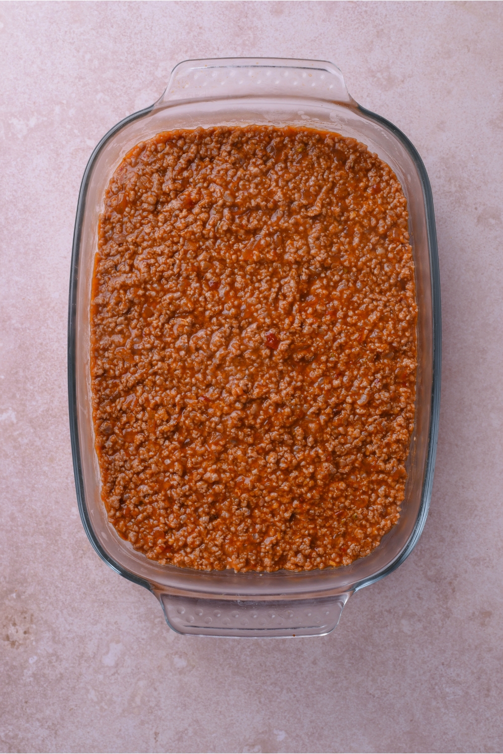 A clear baking dish filled with cooked ground beef in red sauce spread into an even layer.
