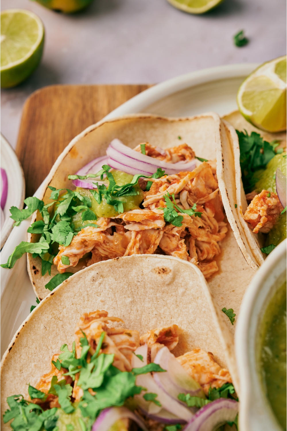 A shredded chicken taco topped with sliced red onion, green salsa, and fresh cilantro. The taco is on a plate with two more tacos, a bowl of salsa, and a lime wedge.