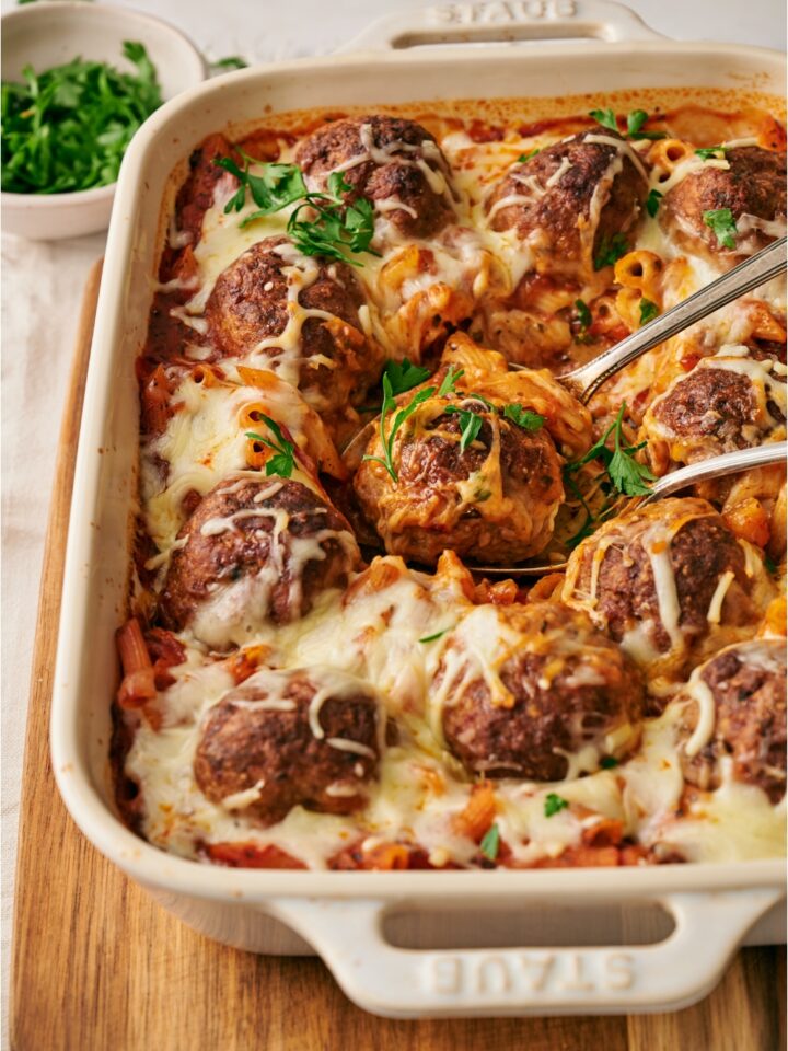 A baking dish filled with pasta and meatballs covered in melted cheese with one meatball being taken out using two spoons.