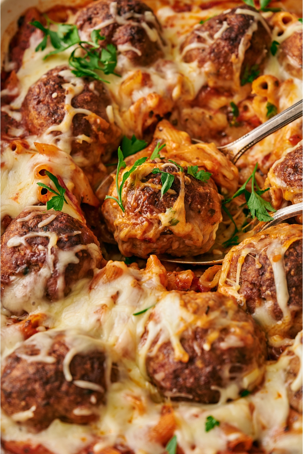Baked pasta and meatballs covered in melted cheese with a single meatball being removed using two spoons.