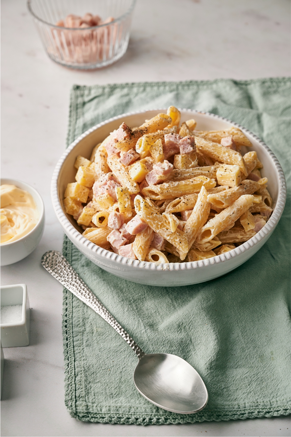 A white bowl of pasta salad with cubed ham, cubed cheese, and black pepper garnished on top, next to a spoon and smaller bowls of cubed ham and mayonnaise.