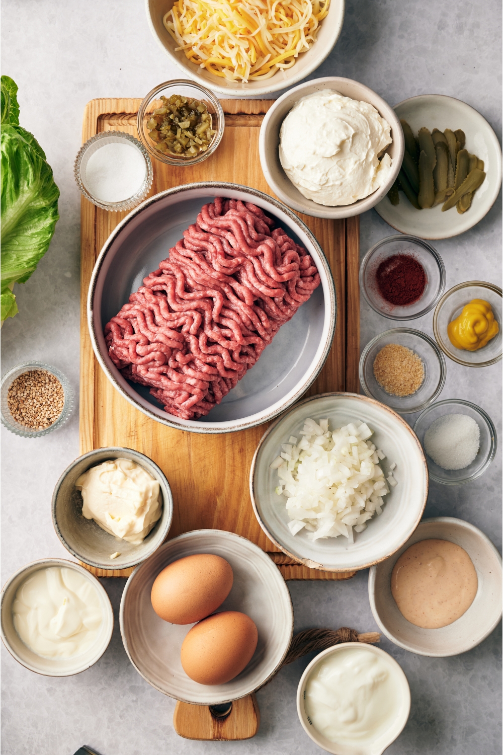 An assortment of ingredients including bowls of raw ground beef, diced onion, two whole eggs, cream cheese, relish, mayonnaise, spices, and a variety of burger toppings.