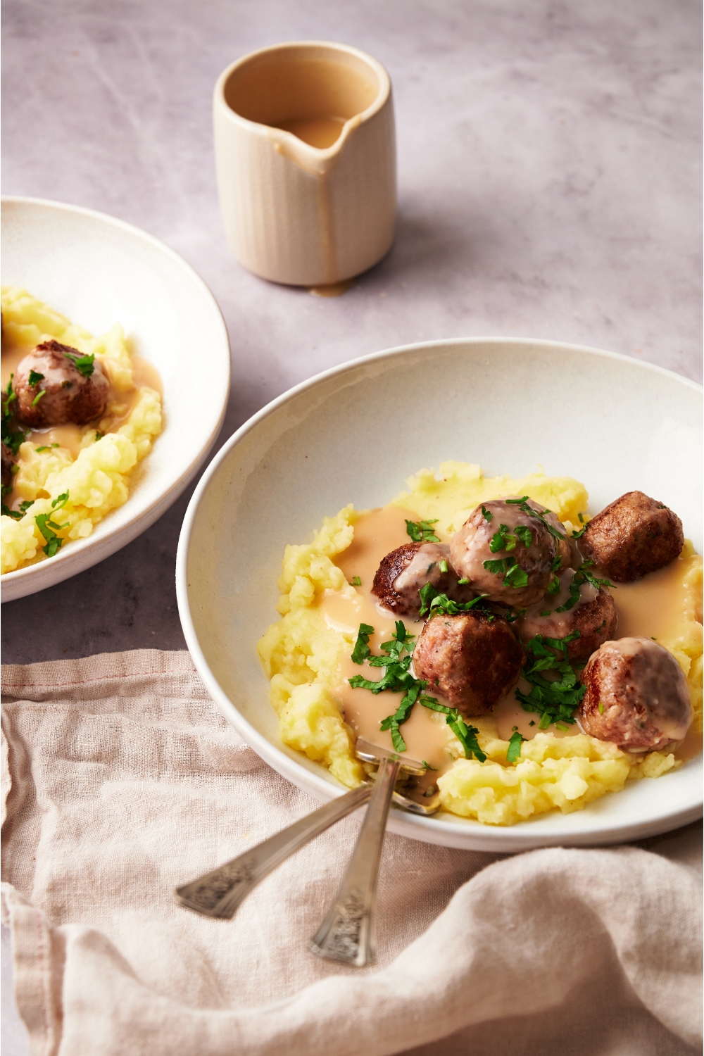 A plate of Swedish meatballs covered in a creamy gravy on a bed of mashed potatoes. There are two forks in the bowl and a garnish of green herbs on top.