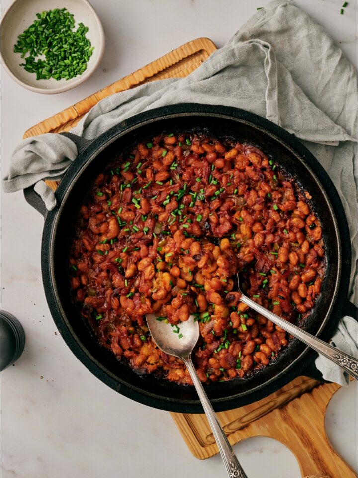 A black skillet atop a wooden board that's filled with freshly baked beans, peppers, onion, garnished with fresh herbs, with two spoons in the skillet.