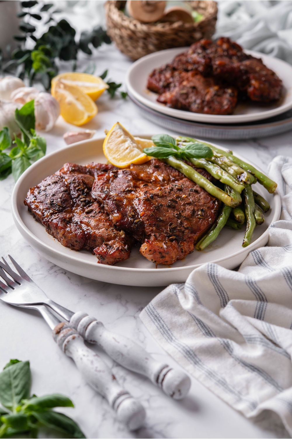 A plate of two seasoned pork steaks layered on top of each other with a side of green beans, a garnish, and two lemon wedges. Next to the plate are two forks and another plate of pork steaks.