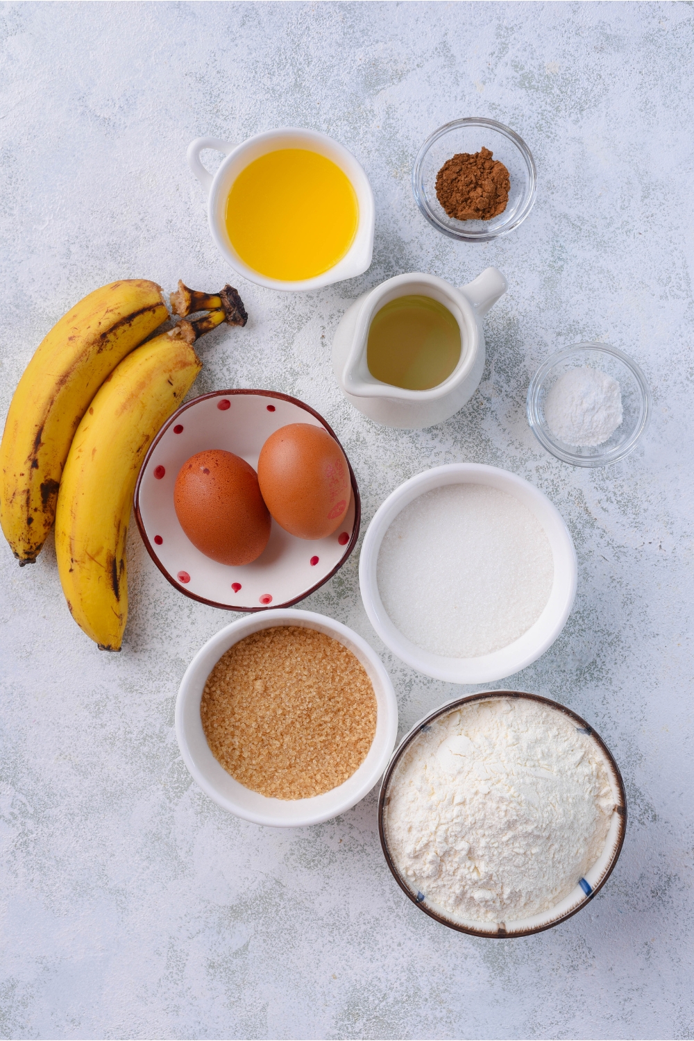An assortment of ingredients including two bananas and bowls of eggs, cinnamon, sugar, brown sugar, flower, baking powder, melted butter, and oil.