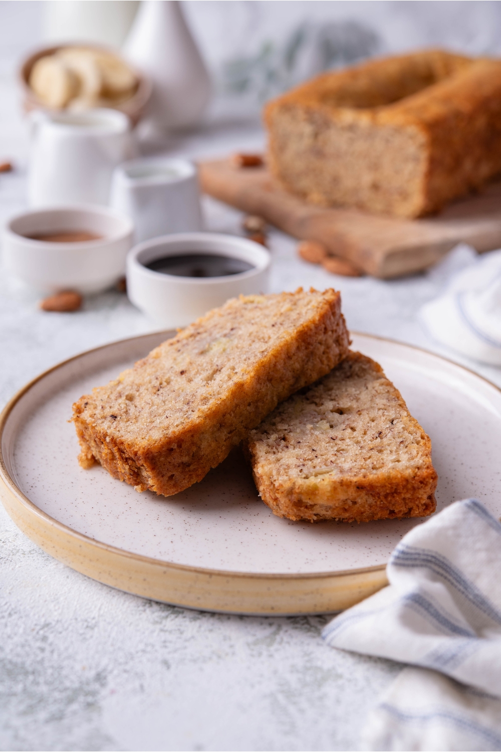 Two slices of banana bread overlapping on a plate with the loaf of banana bread in the background next to bowls of jam.