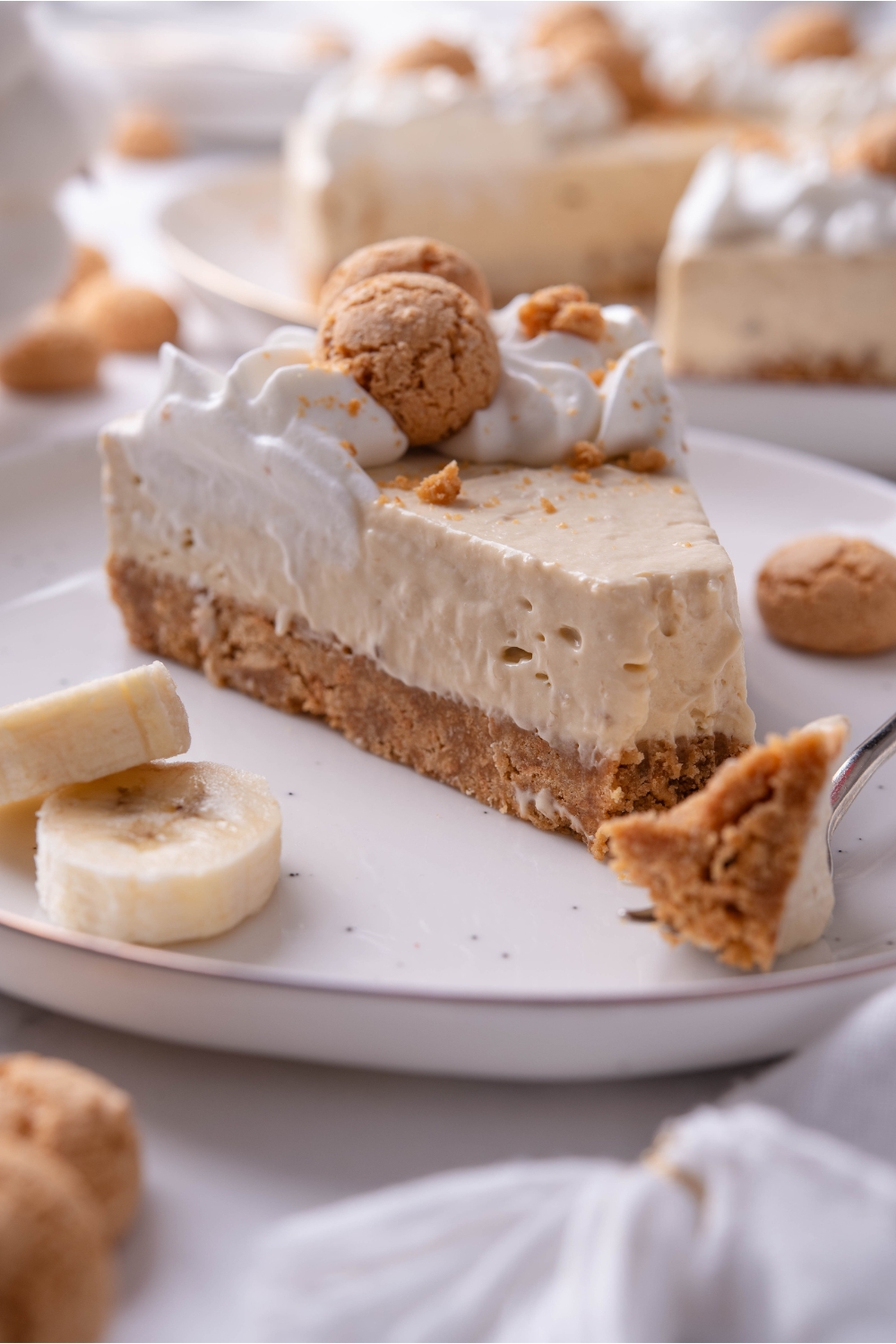 A serving of banana cheesecake on a plate with a bite taken out using a fork that is on the plate. The cheesecake has a cookie crust and is topped with whipped cream and mini cookies.