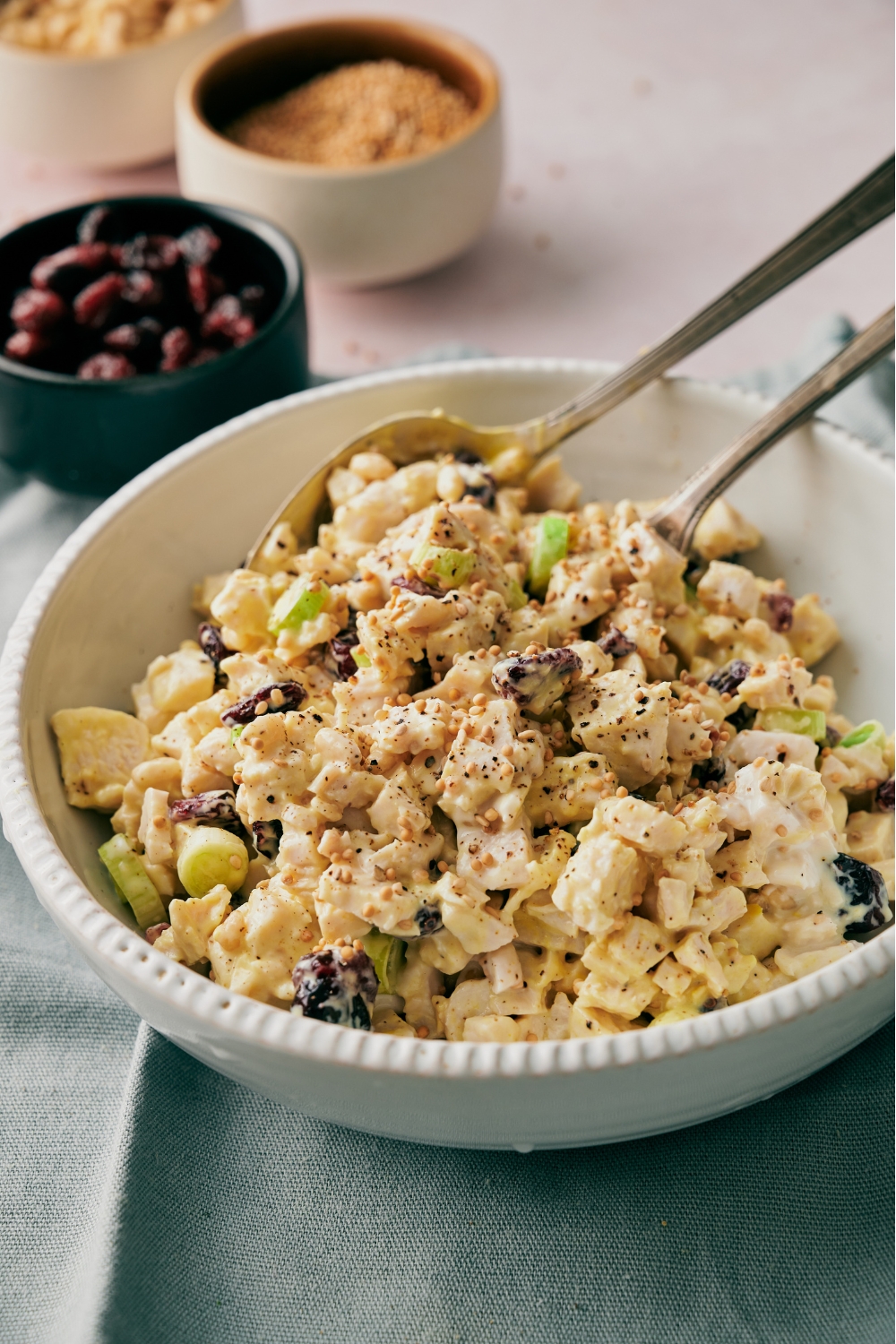 A bowl of chicken salad with diced celery, dried cranberries, and a mustard dressing with black pepper on top. There are two spoons in the bowl.