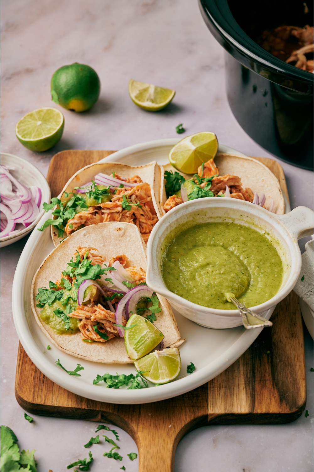 A plate with three shredded chicken tacos topped with sliced red onion and cilantro with a bowl of green salsa and lime wedges on the plate.
