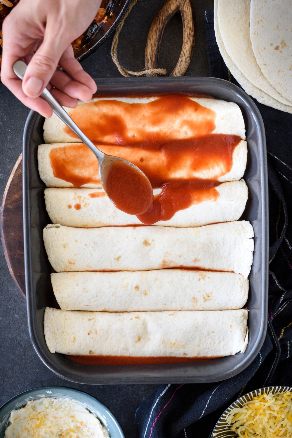 A hand spooning red sauce over top a baking dish filled with rolled tortillas.