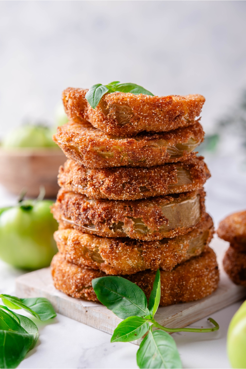 Six breaded and fried green tomatoes stacked evenly on top of each other with a garnish of green herbs on top and fresh green tomatoes surrounding the stack.
