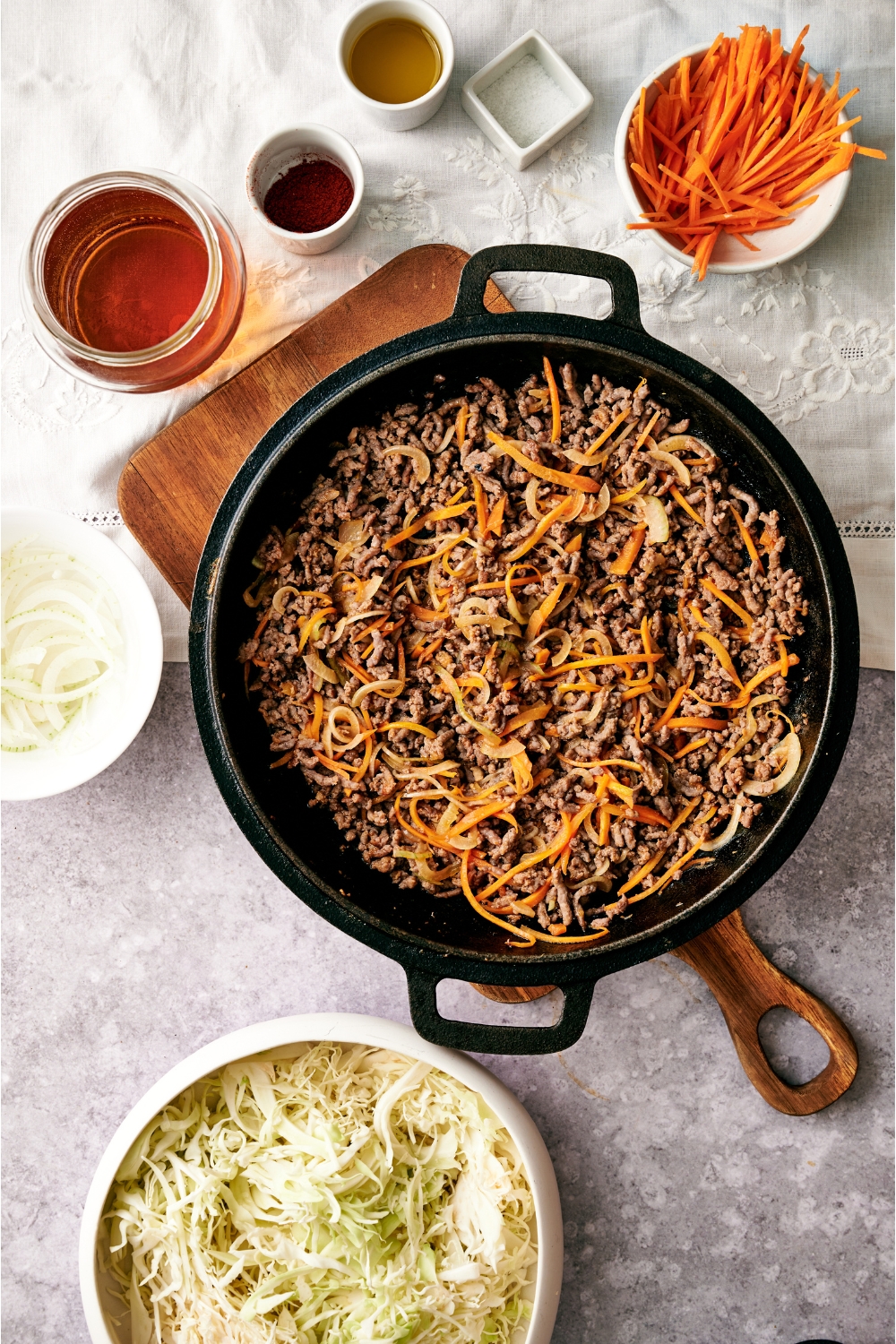 A black skillet filled with cooked ground beef, thinly sliced carrot and onion all mixed together. The skillet is on a wooden board and is surrounded by an assortment of ingredients.