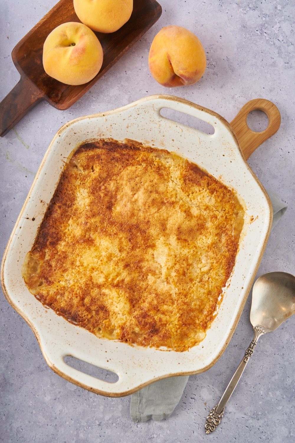 Square baking dish filled with freshly baked peach cobbler covered in a golden brown crust with a serving spoon next to it.