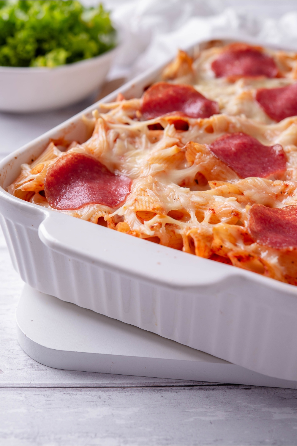 A white baking dish filled with freshly baked pepperoni pasta casserole that's topped with thinly sliced pepperoni and melted cheese.