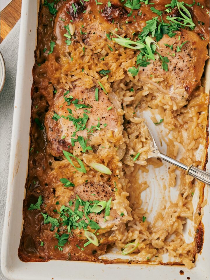 A baking dish filled with freshly baked pork chops and rice in a seasoned cream sauce with green onions on top. A serving is missing from the baking dish and there is a serving spoon in the dish.