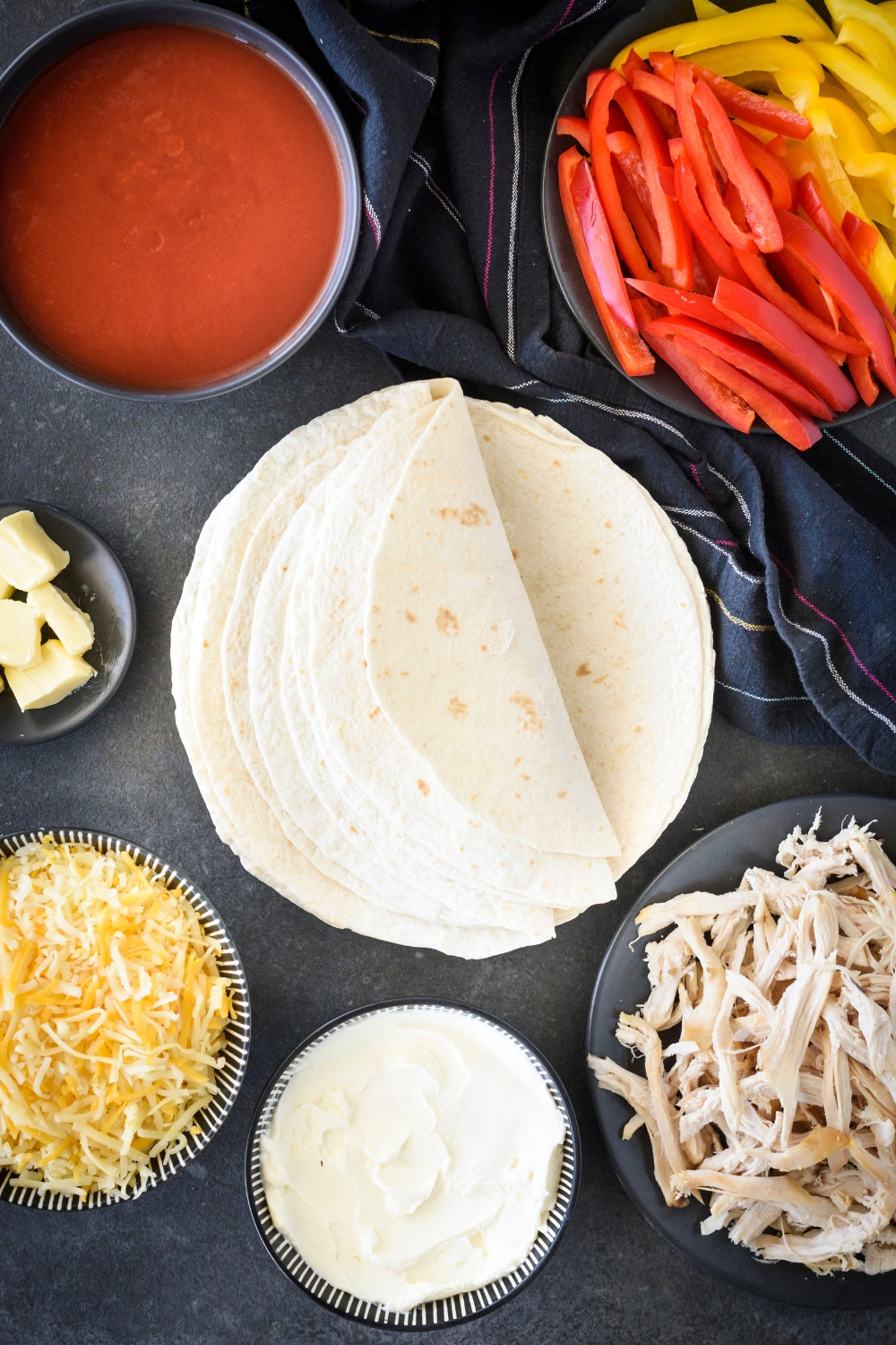 An assortment of ingredients including a pile of flour tortillas surrounded by bowls of enchilada sauce, shredded chicken, shredded cheese, cream cheese, and sliced bell peppers.