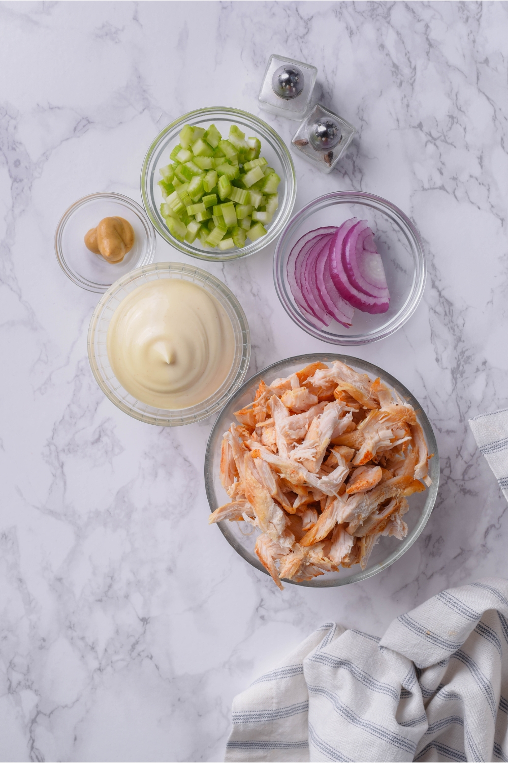 An assortment of ingredients including bowls of shredded seasoned chicken, sliced red onion, mayonnaise, mustard, diced celery, and mini salt and pepper shakers.