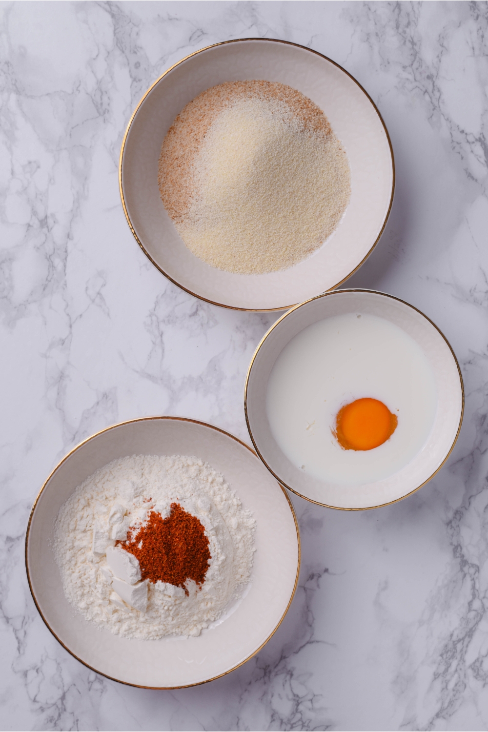 Three bowls, one with milk and an egg, one with flour and paprika, and one with a flour mixture.