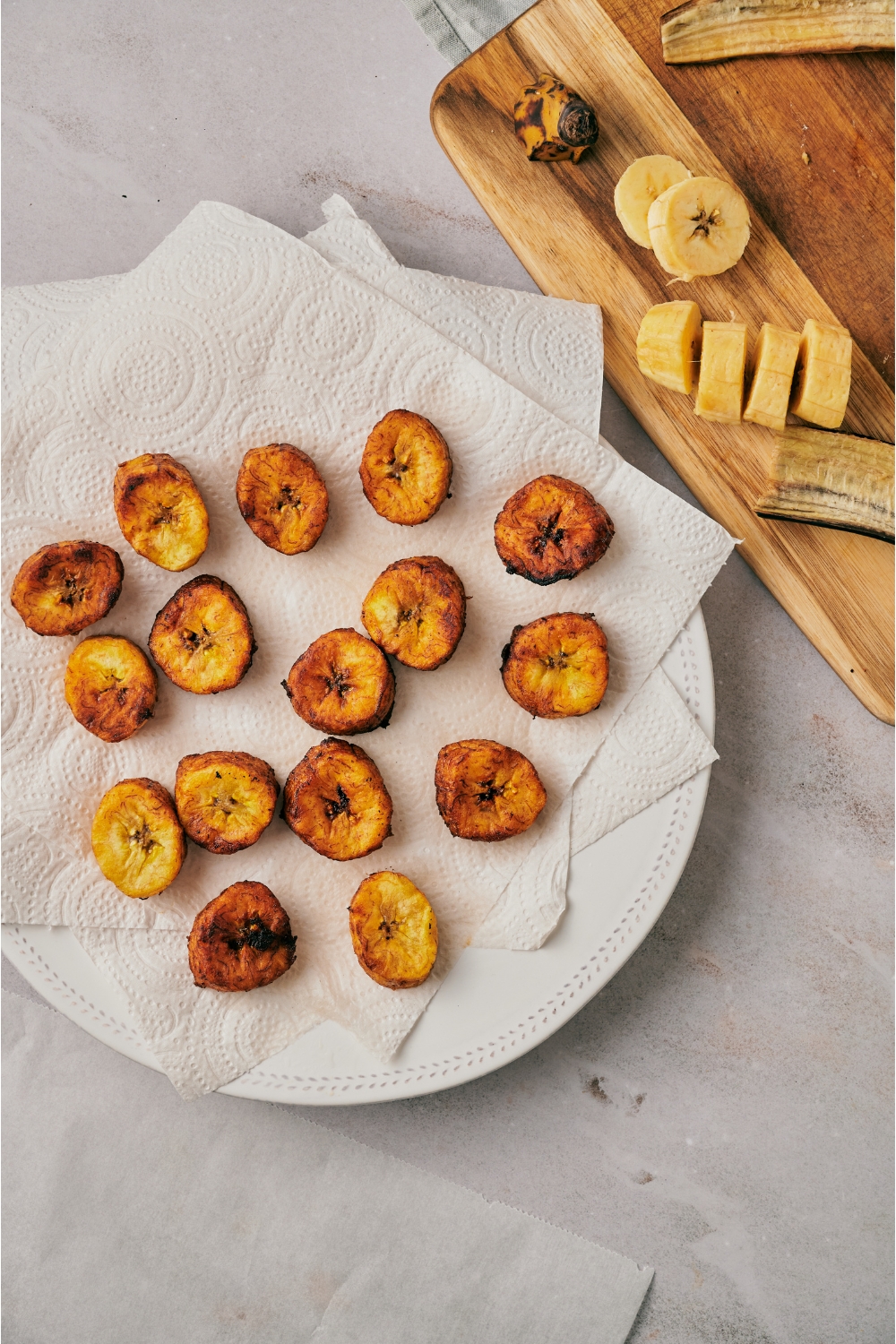 Golden brown plantain slices on a plate lined with three paper towels next to a cutting board with plantain slices and old plantain peels on it.