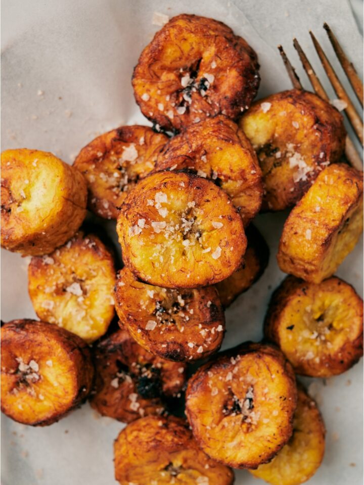 Close up of golden brown deep fried plantain slices covered in coarse sea salt on a plate lined with paper towels. There is a fork on the plate.