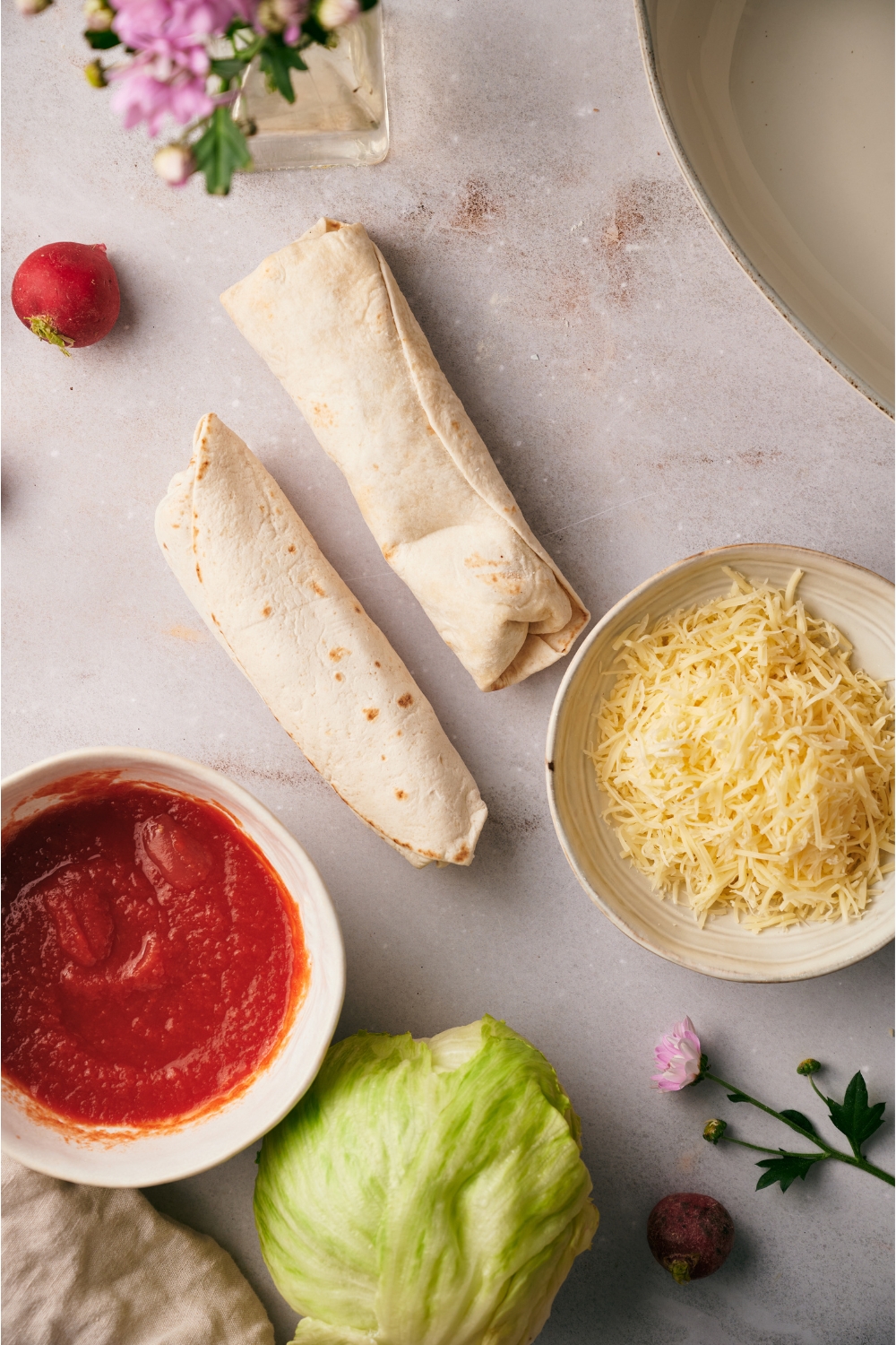 An assortment of ingredients including two frozen burritos, a head of iceberg lettuce, a bowl of shredded cheese, and a bowl of red sauce.