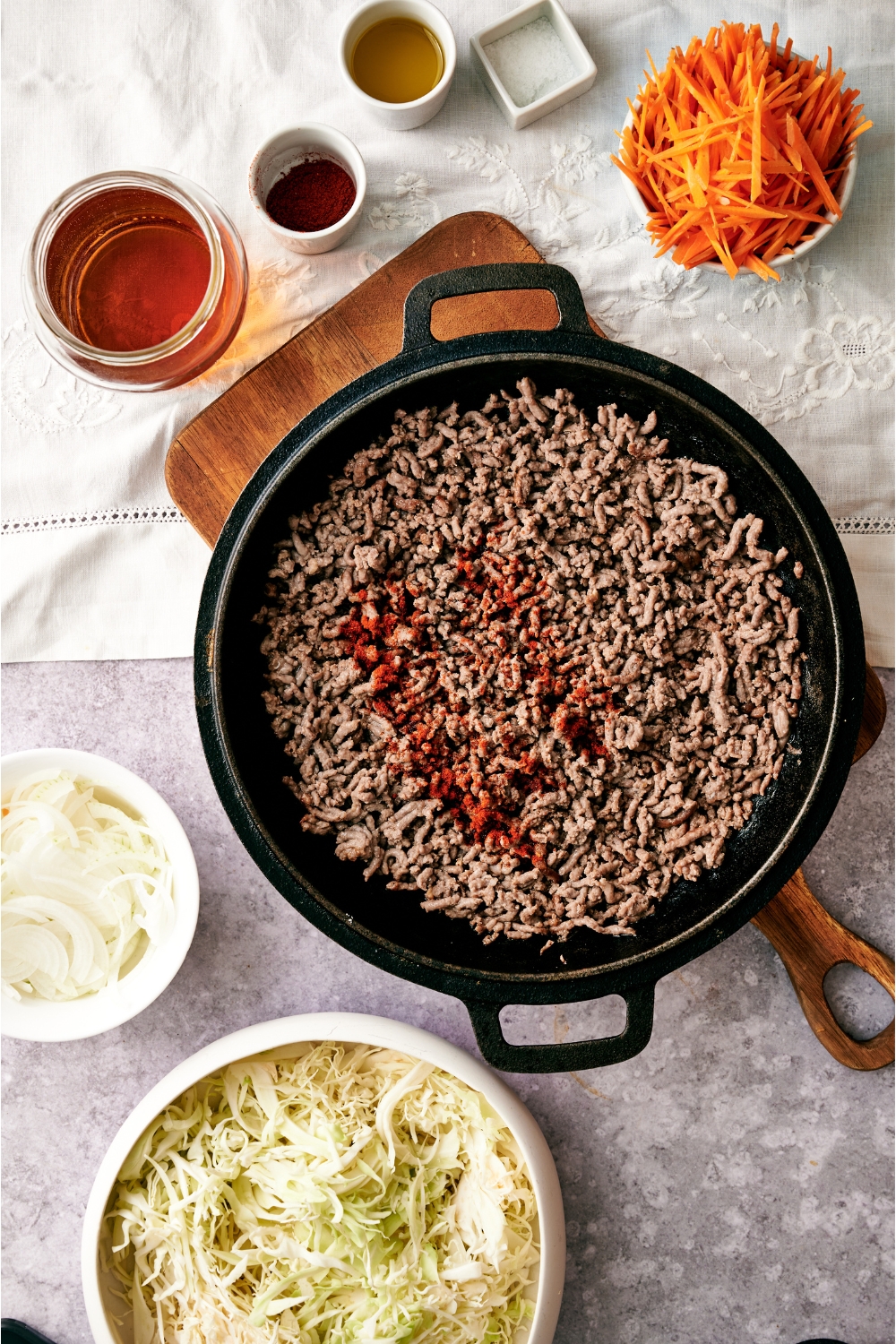 A black skillet filled with cooked ground beef and a sprinkle of paprika on top. The skillet is on a wooden board and is surrounded by an assortment of ingredients.