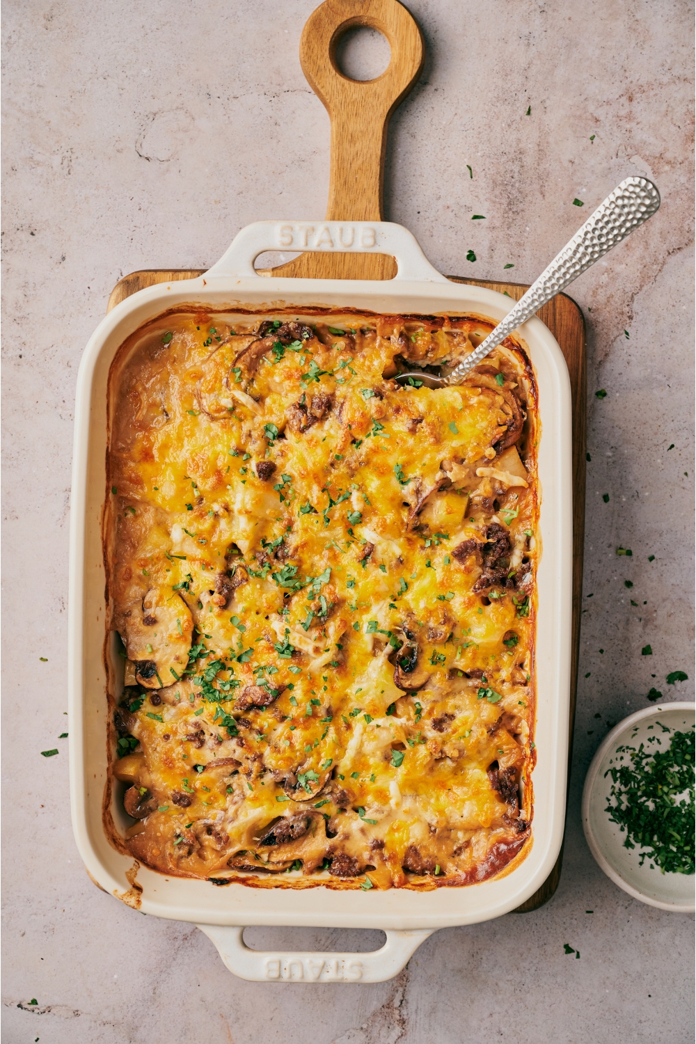 A white baking dish on a wooden board filled with freshly casserole with sliced mushrooms, ground beef, and a layer of melted cheese on top, garnished with fresh parsley.