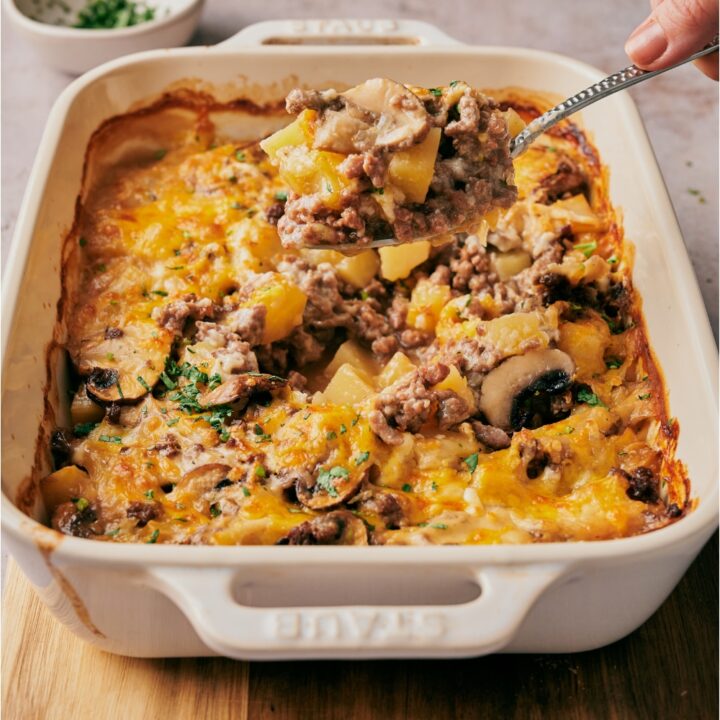 A hand scooping a serving of casserole out of its baking dish. The casserole has ground beef, sliced mushrooms, diced potato, and a layer of melted cheese on top.