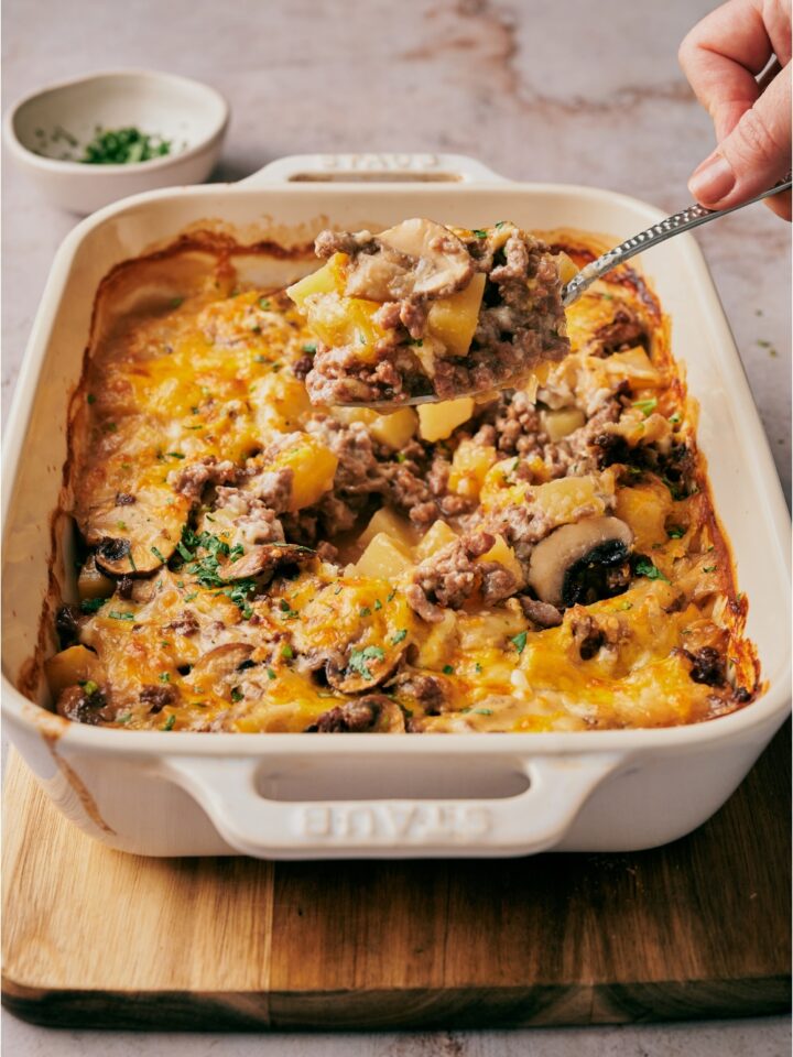 A hand scooping a serving of casserole out of its baking dish. The casserole has ground beef, sliced mushrooms, diced potato, and a layer of melted cheese on top.