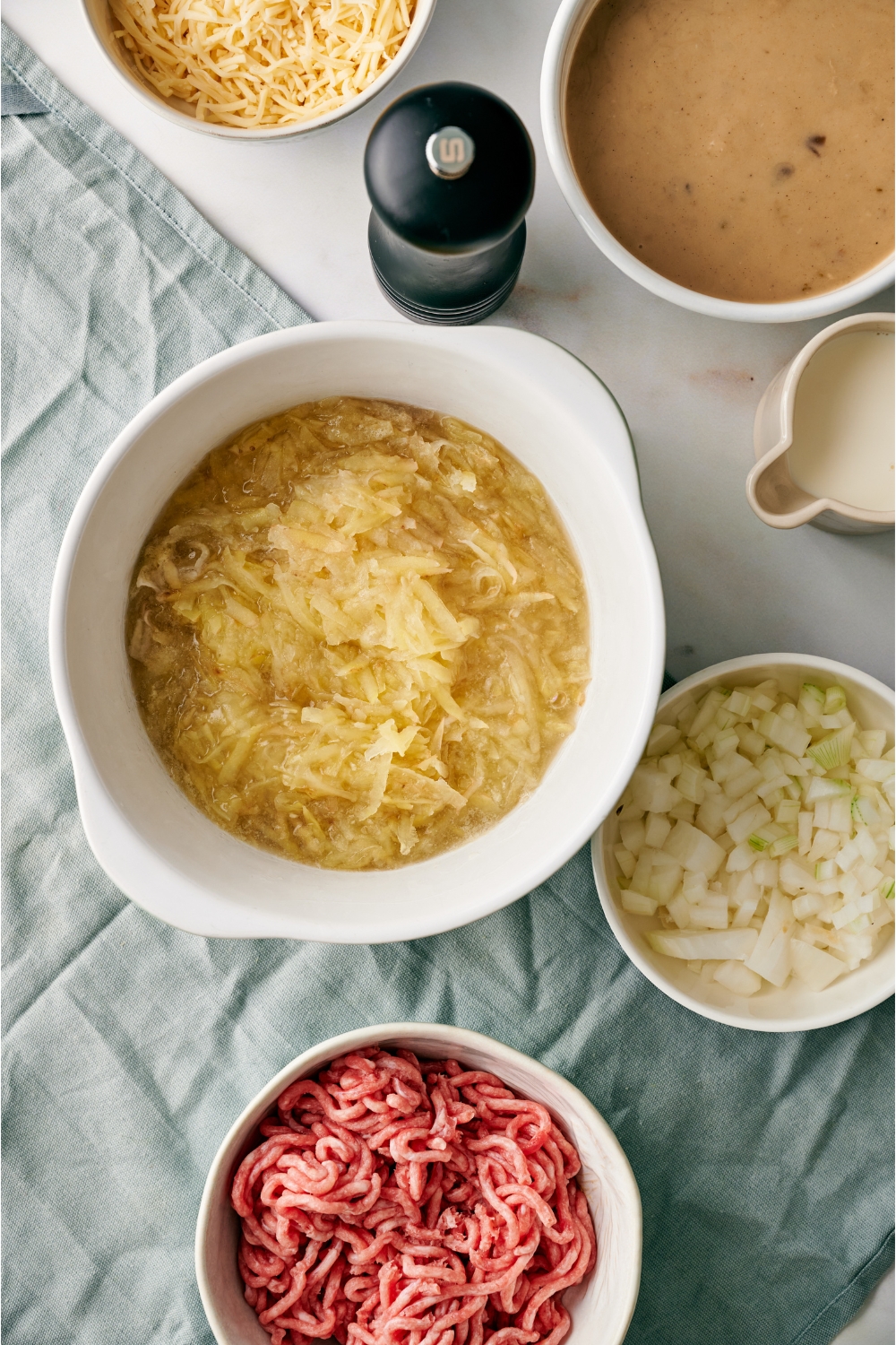 A bowl of thawed hashbrowns next to a black pepper shaker and bowls of raw ground beef, diced onion, cream of mushroom soup, and a pitcher of milk.