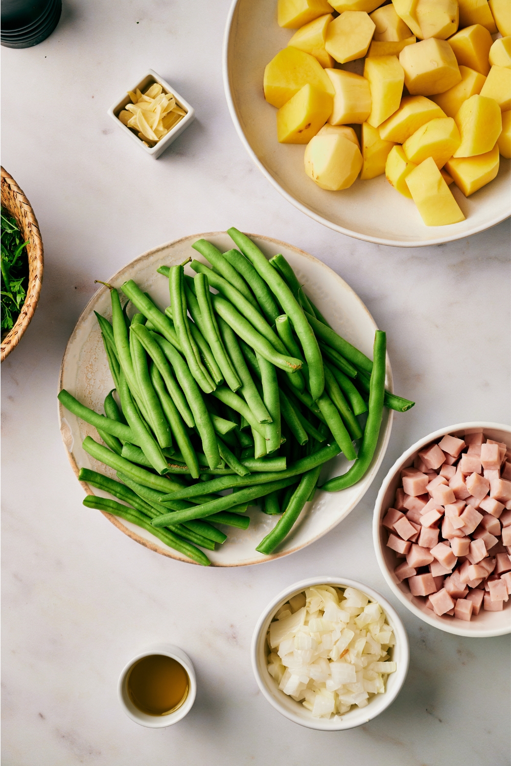 A countertop with ingredients like green beans, onions, ham, potatoes, olive oil, and garlic in separate bowls.