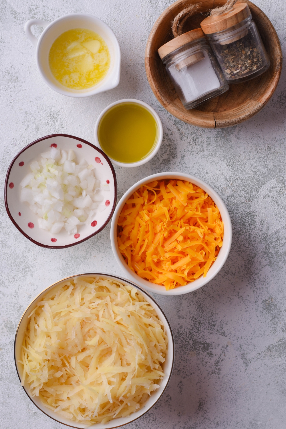 A countertop with separate bowls containing salt and pepper, melted butter, oil, diced onions, shredded carrots, and shredded cheese.