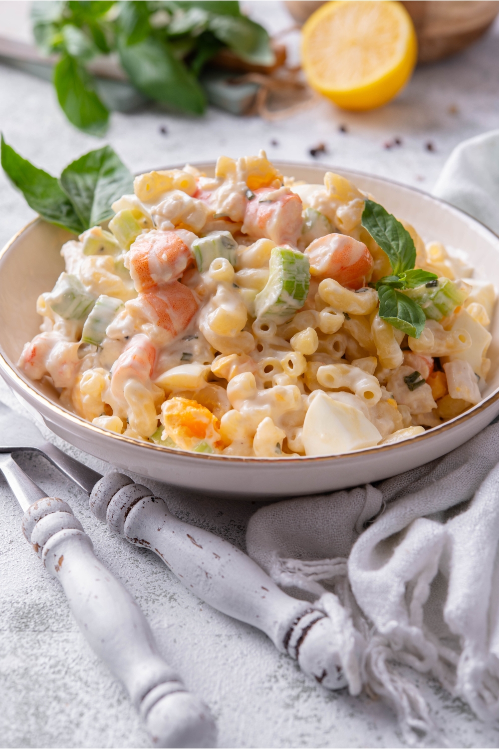 A bowl of seafood pasta salad with macaroni noodles, creamy dressing, and chunks of celery, hard-boiled eggs, and shrimp. The pasta salad is garnished with basil and two forks are next to the bowl.