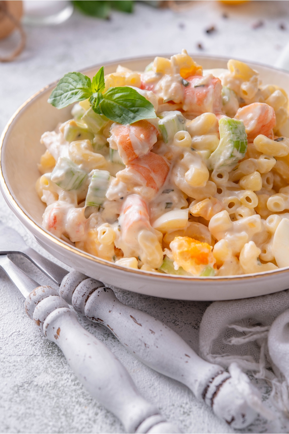 A bowl of seafood pasta salad with macaroni noodles, creamy dressing, and chunks of celery, hard-boiled eggs, and shrimp. The pasta salad is garnished with basil and two forks are next to the bowl.
