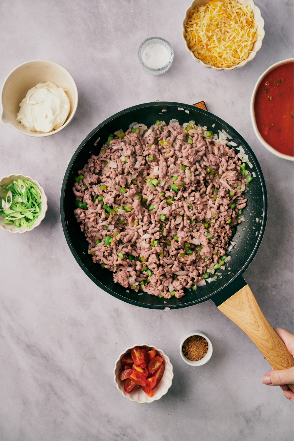 A skillet filled with cooked ground beef, diced onion, and diced green peppers. The skillet is surrounded by an assortment of ingredients.