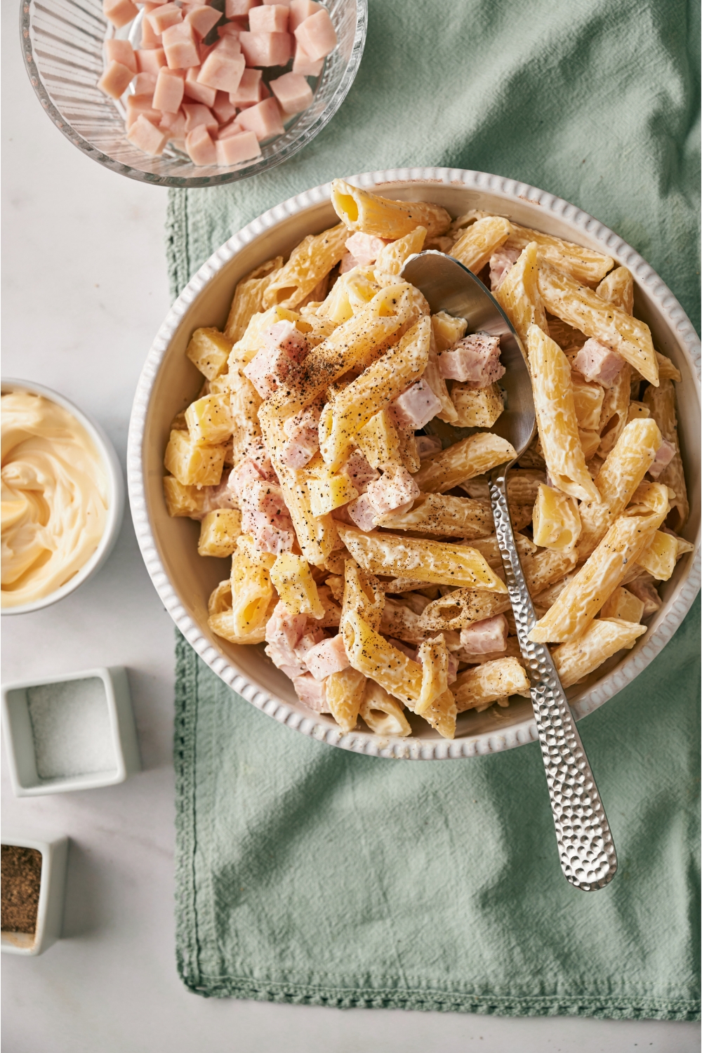 A white bowl filled with pasta salad, with penne pasta, cubed ham, cubed cheese, and black pepper, all tossed in a creamy dressing using a spoon in the bowl.